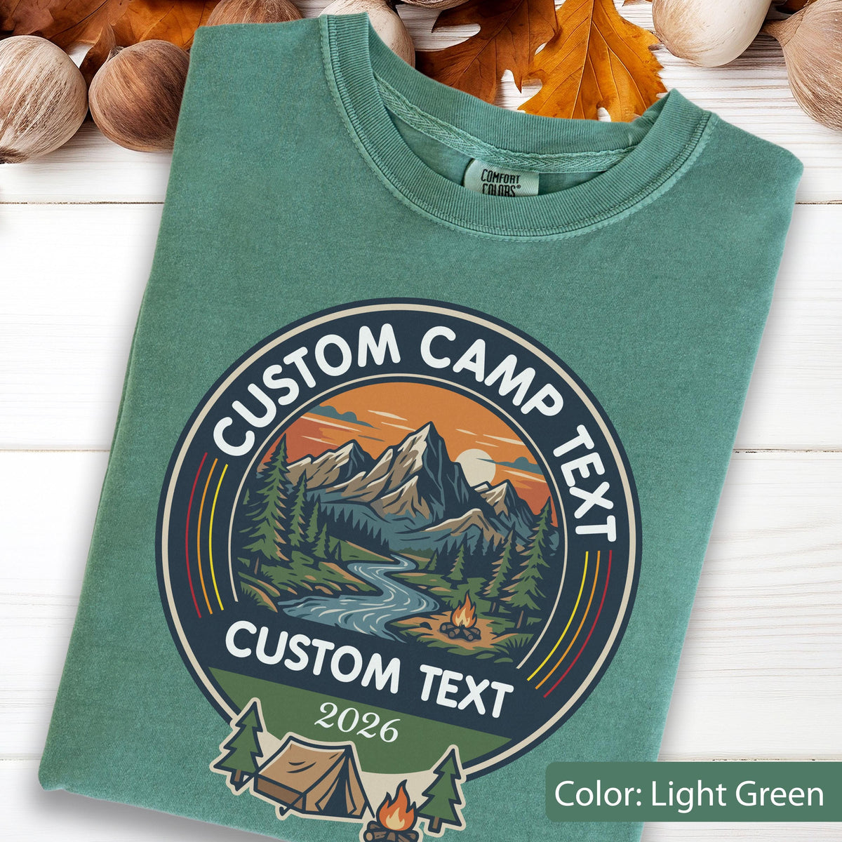 May include: A light green t-shirt featuring a circular graphic with a mountain landscape, a river, a campfire, and a tent. The text reads CUSTOM CAMP TEXT, CUSTOM TEXT, and 2026. The shirt has a Comfort Colors label at the inner neckline.