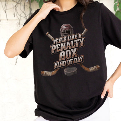 May include: A black short-sleeved t-shirt featuring a graphic of a hockey helmet, two crossed hockey sticks, and a puck. The text reads, "FEELS LIKE A PENALTY BOX KIND OF DAY" in a distressed, weathered font style centered on the chest area.