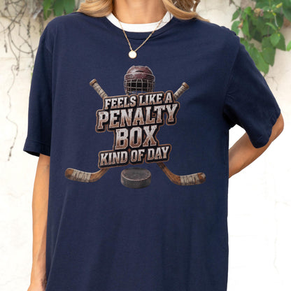 May include: A navy blue t-shirt featuring a graphic of a hockey helmet, two crossed hockey sticks, and a puck. The text reads, FEELS LIKE A PENALTY BOX KIND OF DAY, in a distressed, block-style font centered on the chest of the garment.