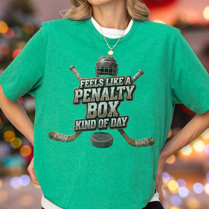 May include: A bright green short-sleeve t-shirt featuring a graphic of a hockey helmet, two crossed hockey sticks, and a puck. The text reads FEELS LIKE A PENALTY BOX KIND OF DAY in a distressed, weathered font centered on the chest area.