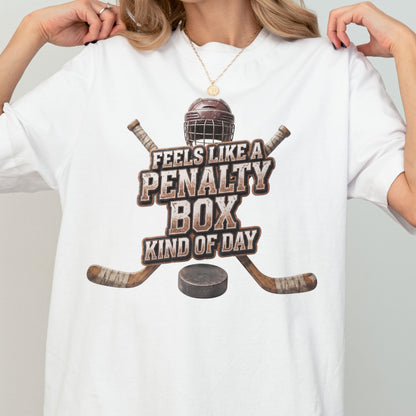 May include: A white t-shirt featuring a graphic of a brown hockey helmet, two crossed wooden hockey sticks, and a black puck. The text reads FEELS LIKE A PENALTY BOX KIND OF DAY in a distressed brown font centered above the hockey puck graphic.
