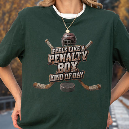 May include: A dark green short-sleeve t-shirt featuring a graphic of a hockey helmet, two crossed hockey sticks, and a puck. The text reads FEELS LIKE A PENALTY BOX KIND OF DAY in a distressed, weathered font centered on the chest of the garment.