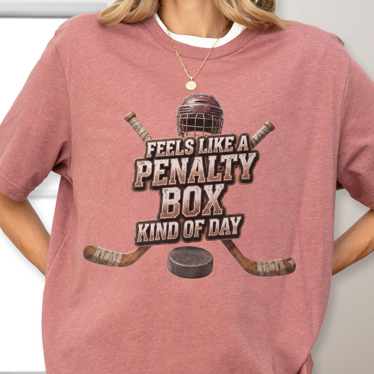 May include: A dusty rose short-sleeve t-shirt featuring a graphic of a hockey helmet, two crossed hockey sticks, and a puck. The text reads FEELS LIKE A PENALTY BOX KIND OF DAY in a distressed, block-style font centered on the chest area.