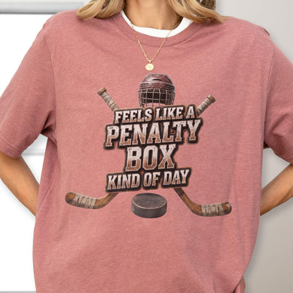 May include: A dusty rose short-sleeve t-shirt featuring a graphic of a hockey helmet, two crossed hockey sticks, and a puck. The text reads FEELS LIKE A PENALTY BOX KIND OF DAY in a distressed, block-style font centered on the chest area.