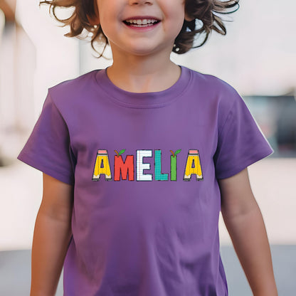 May include: A purple short-sleeved t-shirt featuring the name AMELIA printed across the chest. Each letter is designed with school-themed patterns, including yellow pencils, red apples, white lined notebook paper, and green grid paper textures.
