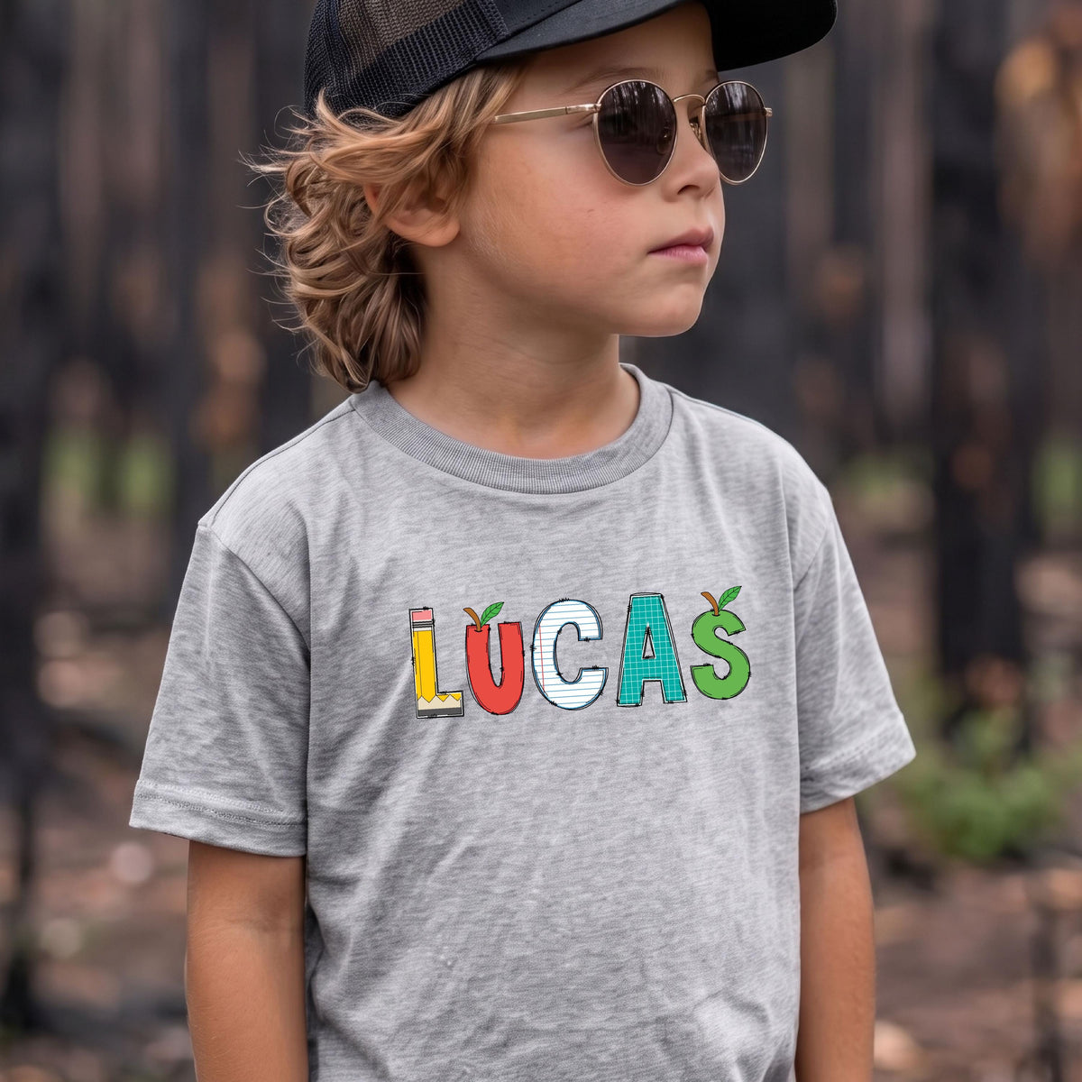 May include: A heather gray short-sleeved t-shirt featuring the name LUCAS printed in colorful, school-themed letters. The L is a yellow pencil, the U is a red apple, the C is lined paper, the A is a teal grid, and the S is a green apple.