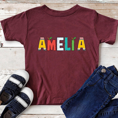 May include: A maroon short-sleeved t-shirt featuring the name AMELIA printed in the center. Each letter is designed with school-themed graphics, including yellow pencils, a red apple, lined notebook paper, and a green grid pattern.