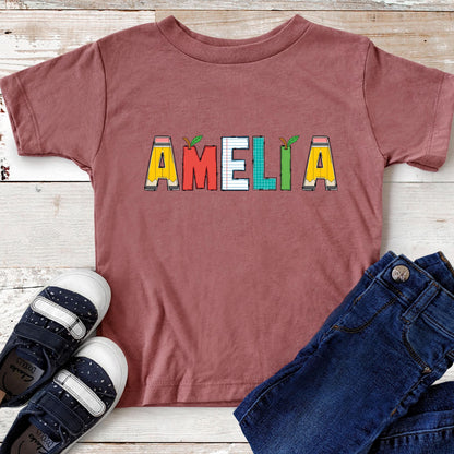 May include: A mauve short-sleeved t-shirt featuring the name AMELIA printed in a school-themed font. Each letter is styled like a different school supply, including pencils, a red apple, notebook paper, and a green grid pattern.