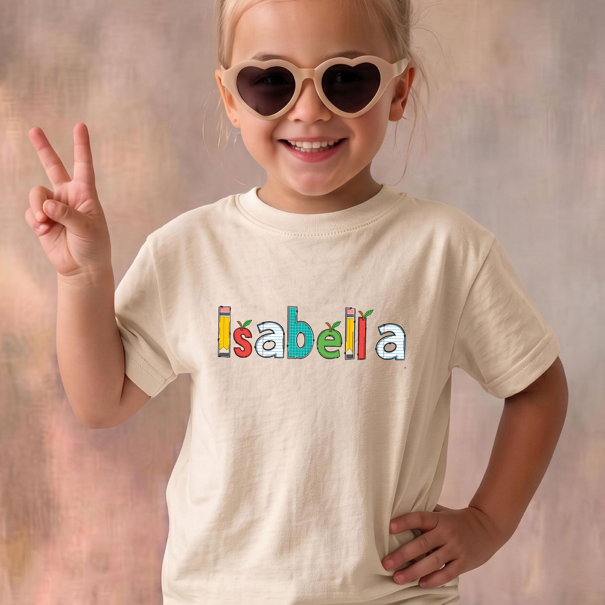 May include: A cream-colored short-sleeve t-shirt featuring the name Isabella printed in a colorful, school-themed font. The letters are designed to look like pencils, apples, and grid paper, creating a playful and educational aesthetic on the chest.