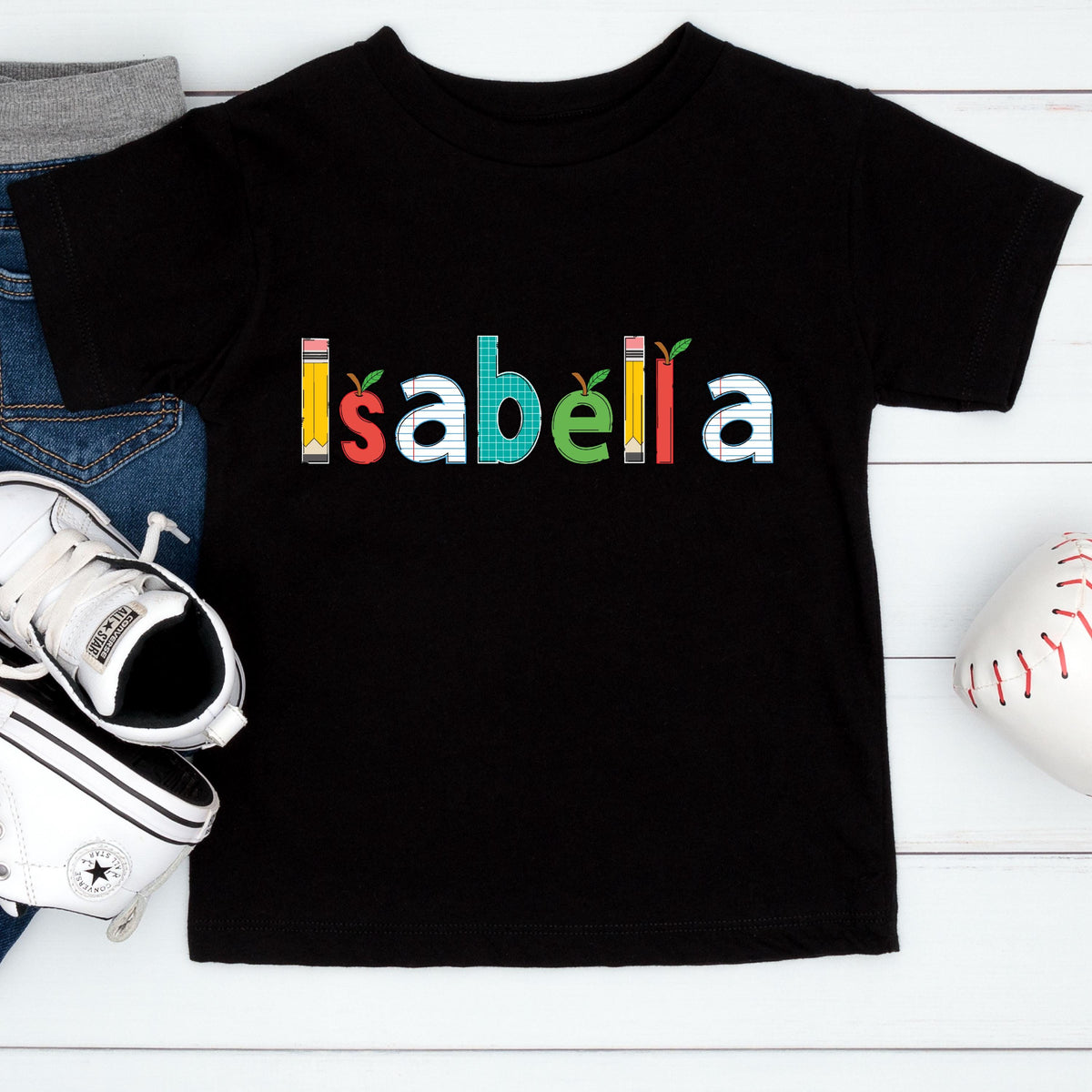 May include: A black short-sleeved t-shirt featuring the name Isabella printed in the center. The letters are stylized with school-themed graphics, including yellow pencils, red and green apples, and blue lined paper patterns integrated into the design.