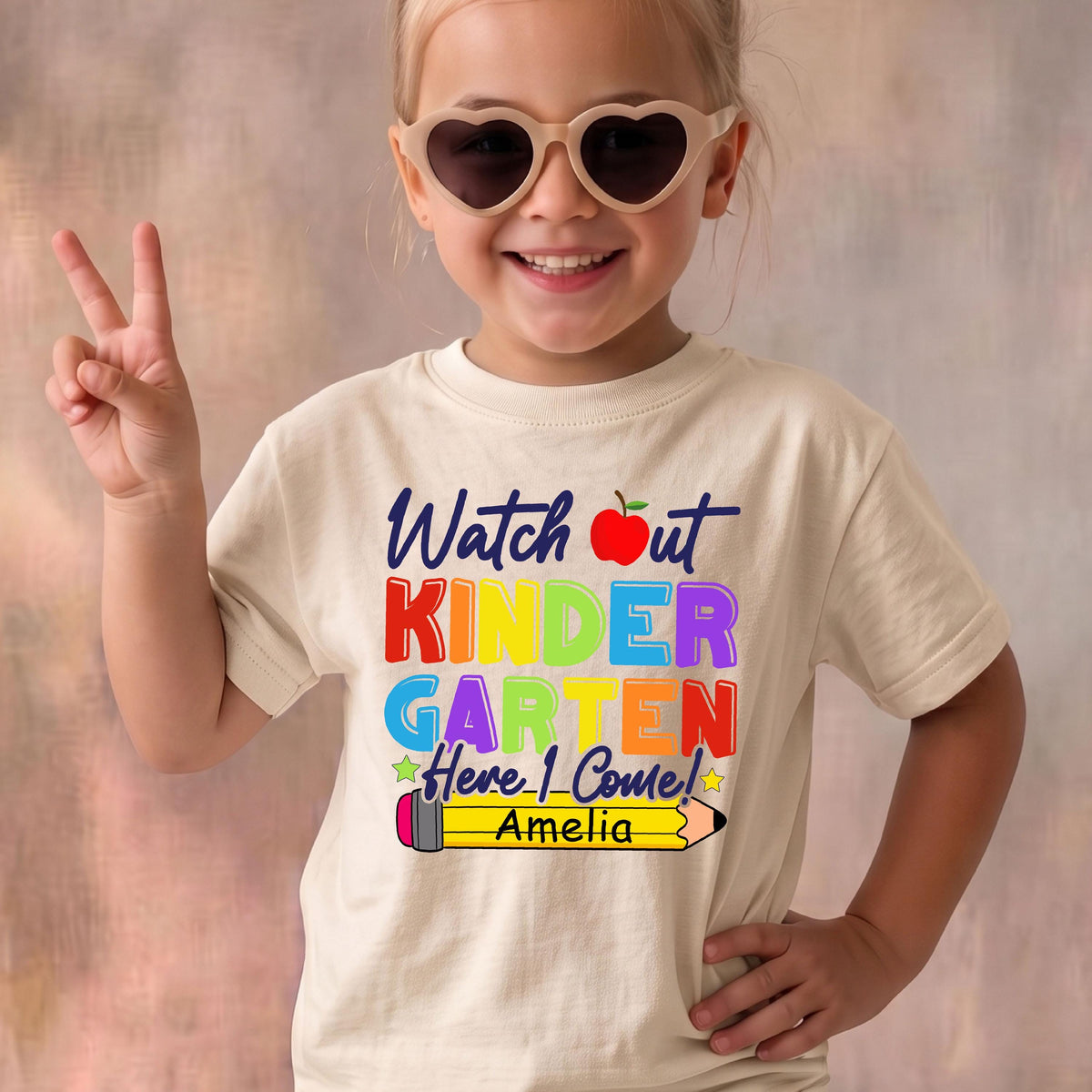 May include: A cream-colored short-sleeve t-shirt featuring the text Watch out KINDERGARTEN Here I Come! in multi-colored letters, a red apple graphic, and a yellow pencil graphic with the name Amelia printed in black on the pencil.