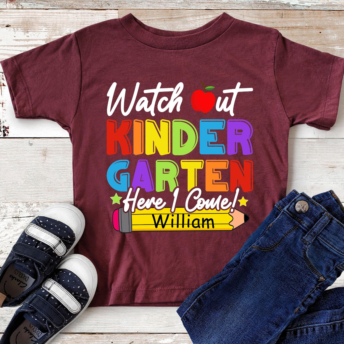 May include: A maroon short-sleeve t-shirt featuring the white text Watch out, a red apple icon, and the colorful text KINDERGARTEN. Below, the text Here I Come! appears above a yellow pencil graphic with the name William printed in black.