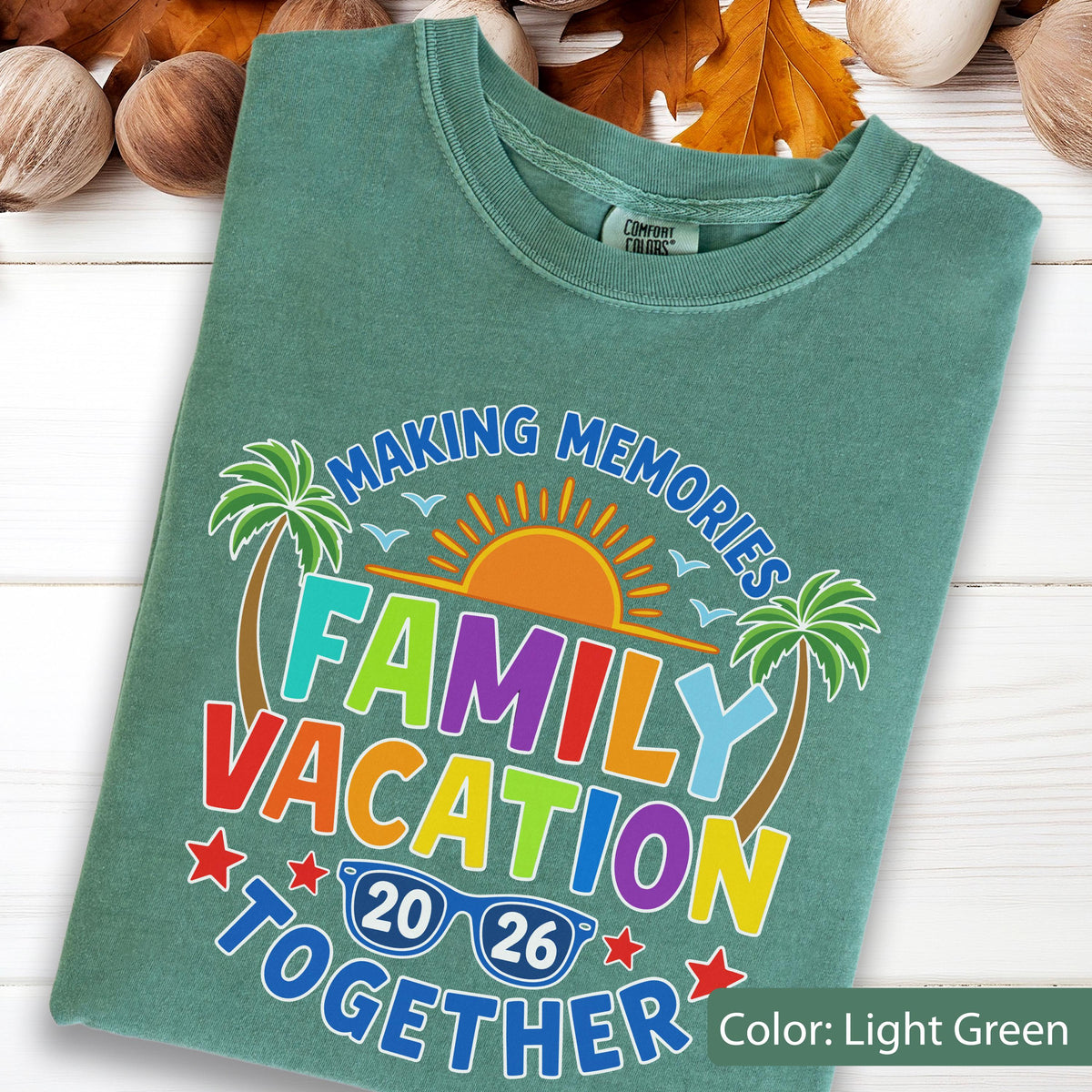 May include: A light green t-shirt featuring a graphic with the text "MAKING MEMORIES FAMILY VACATION TOGETHER 2026". The design includes a bright orange sun, two green palm trees, small blue birds, and a pair of blue sunglasses with the year 2026.