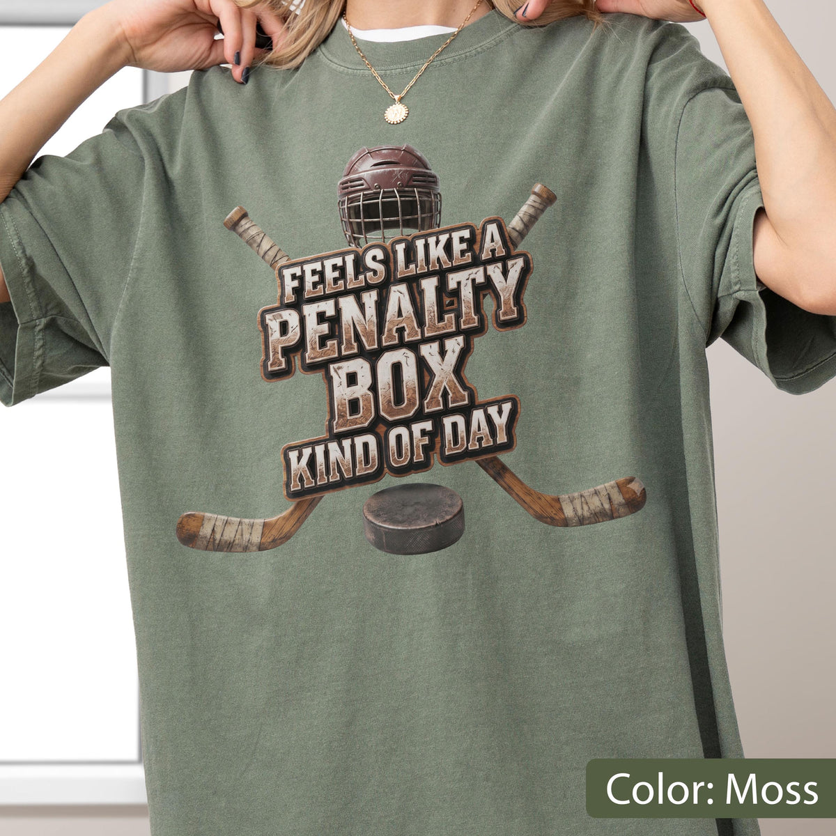 May include: A moss green short-sleeve t-shirt featuring a graphic of a hockey helmet, two crossed hockey sticks, and a puck. The text reads, "FEELS LIKE A PENALTY BOX KIND OF DAY," printed in a distressed, block-style font across the center chest.