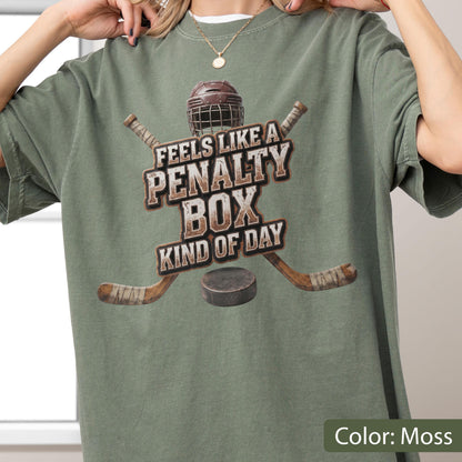 May include: A moss green short-sleeve t-shirt featuring a graphic of a hockey helmet, two crossed hockey sticks, and a puck. The text reads, "FEELS LIKE A PENALTY BOX KIND OF DAY," printed in a distressed, block-style font across the center chest.