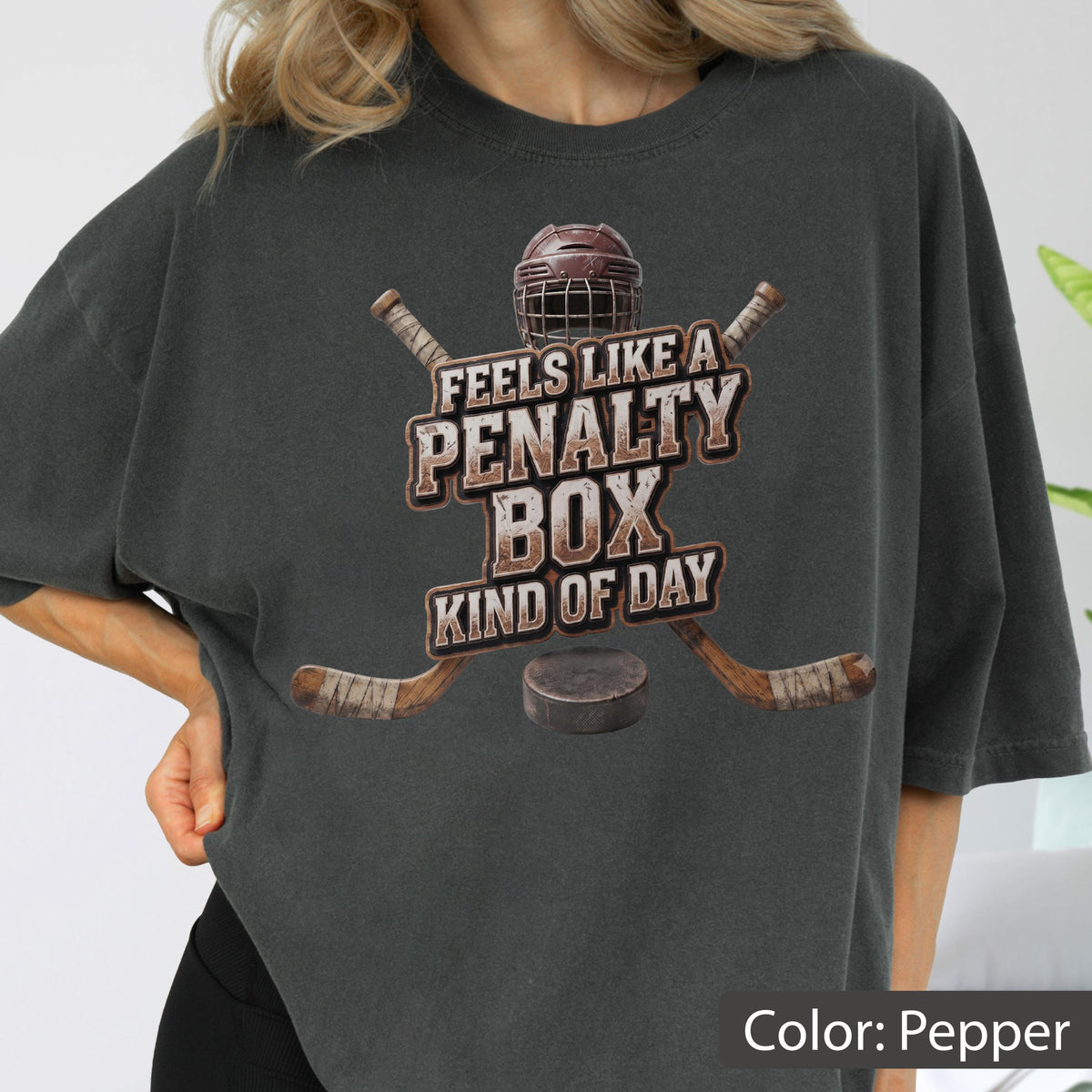 May include: A charcoal gray t-shirt featuring a graphic of a hockey helmet, two crossed hockey sticks, and a puck. The text reads FEELS LIKE A PENALTY BOX KIND OF DAY in a distressed, block-style font centered above the hockey equipment.