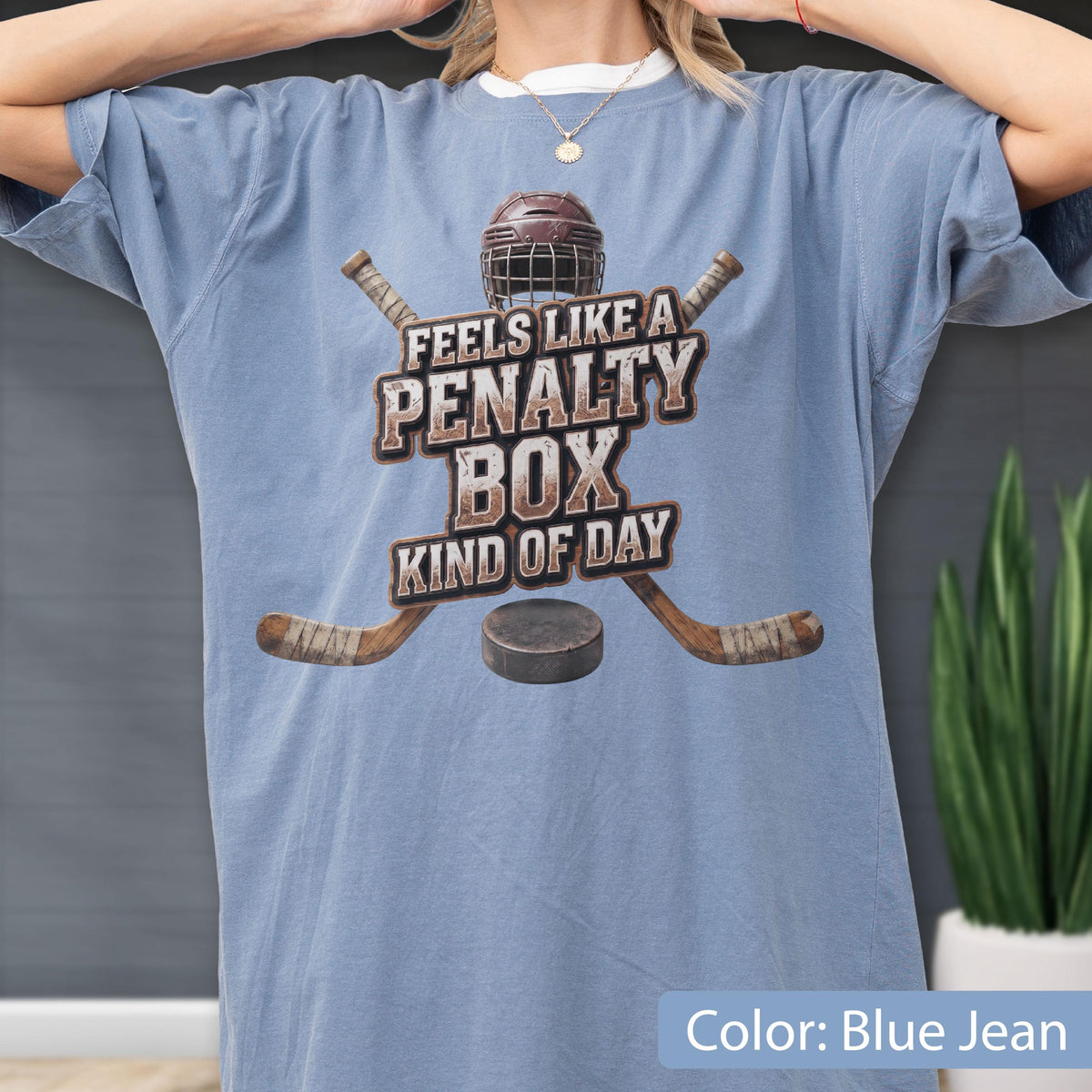 May include: A blue jean colored t-shirt featuring a graphic of a hockey helmet, two crossed hockey sticks, and a puck. The text reads, FEELS LIKE A PENALTY BOX KIND OF DAY, in a distressed brown font centered on the chest of the garment.