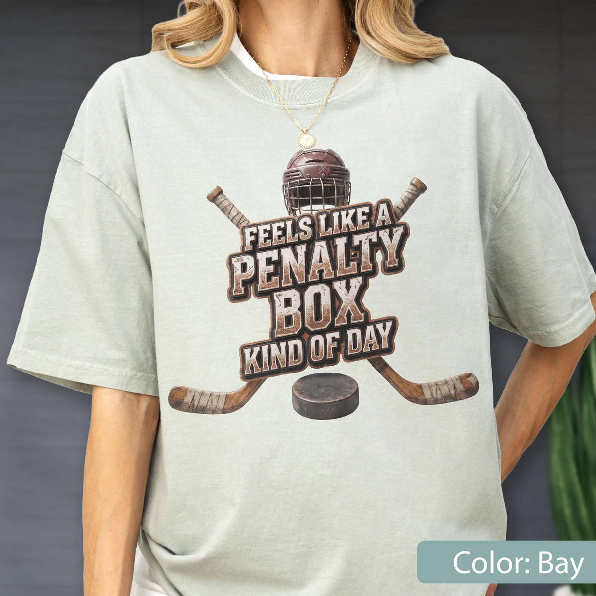 May include: A light sage green short-sleeve t-shirt featuring a graphic of a hockey helmet, two crossed hockey sticks, and a puck. The text reads "FEELS LIKE A PENALTY BOX KIND OF DAY" in a distressed brown font centered on the chest area.