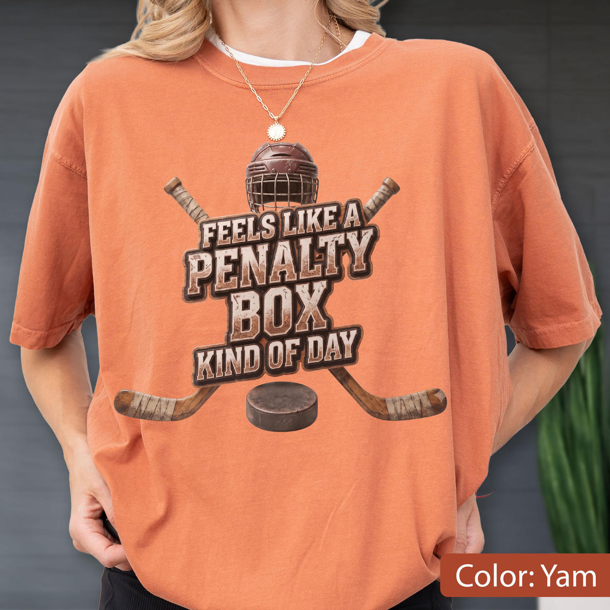 May include: An orange short-sleeve t-shirt featuring a graphic of a hockey helmet, two crossed hockey sticks, and a puck. The text reads FEELS LIKE A PENALTY BOX KIND OF DAY in a distressed brown font centered on the chest of the garment.
