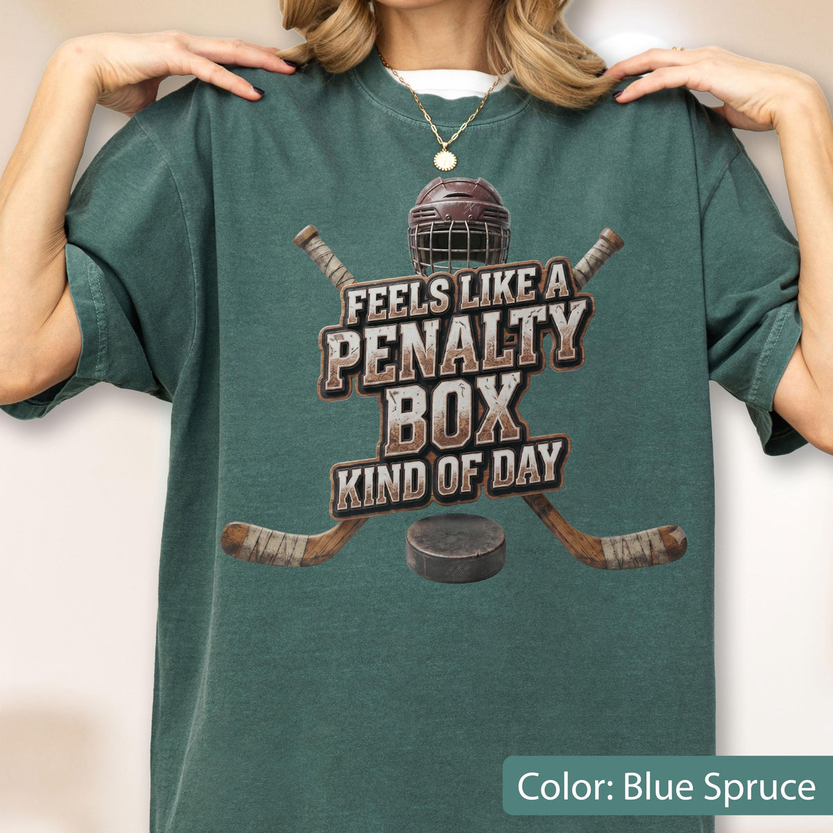 May include: A blue spruce t-shirt featuring a graphic of a hockey helmet, two crossed hockey sticks, and a puck. The text reads FEELS LIKE A PENALTY BOX KIND OF DAY in a distressed font. The shirt has a crew neckline and short sleeves.