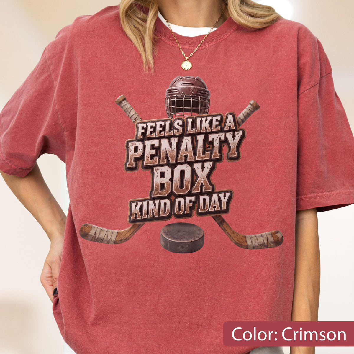 May include: A crimson short-sleeve t-shirt featuring a graphic of a hockey helmet, two crossed hockey sticks, and a puck. The text reads FEELS LIKE A PENALTY BOX KIND OF DAY in a distressed brown font centered on the chest of the garment.