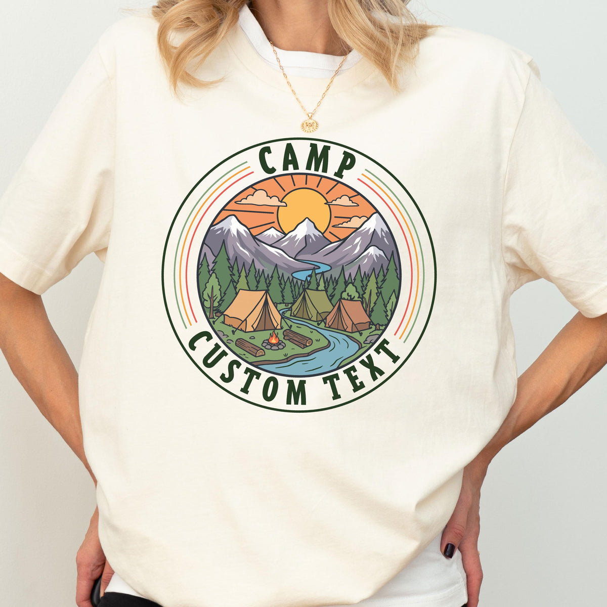 May include: A cream-colored t-shirt featuring a circular graphic of a mountain landscape with tents, a campfire, and a river. The text CAMP and CUSTOM TEXT are printed in dark green within the circular border, surrounding the outdoor scene.