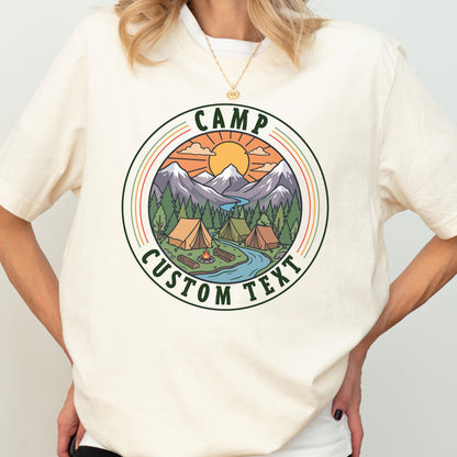 May include: A cream-colored t-shirt featuring a circular graphic of a mountain landscape with tents, a campfire, and a river. The text CAMP and CUSTOM TEXT are printed in dark green within the circular border, surrounding the outdoor scene.