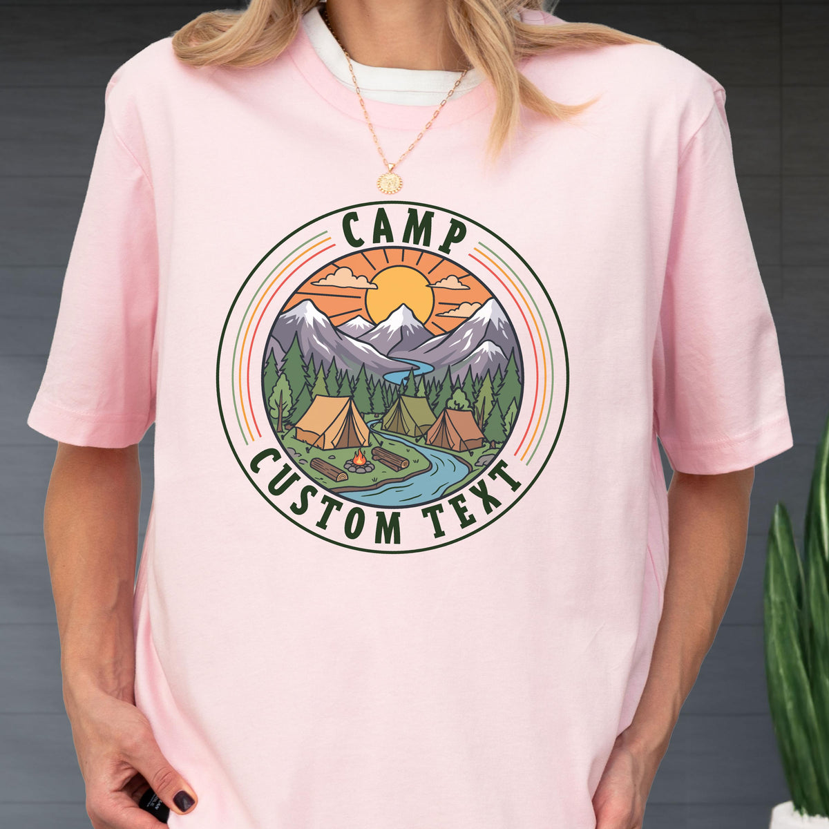 May include: A light pink t-shirt featuring a circular graphic with a mountain landscape, pine trees, tents, a campfire, and a river under a sun. The text CAMP is printed at the top and CUSTOM TEXT is printed at the bottom of the circular design.