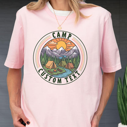 May include: A light pink t-shirt featuring a circular graphic with a mountain landscape, pine trees, tents, a campfire, and a river under a sun. The text CAMP is printed at the top and CUSTOM TEXT is printed at the bottom of the circular design.