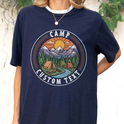 May include: A navy blue t-shirt featuring a circular graphic with a mountain landscape, a river, pine trees, tents, and a campfire. The text "CAMP" is arched at the top and "CUSTOM TEXT" is arched at the bottom within the circular border.
