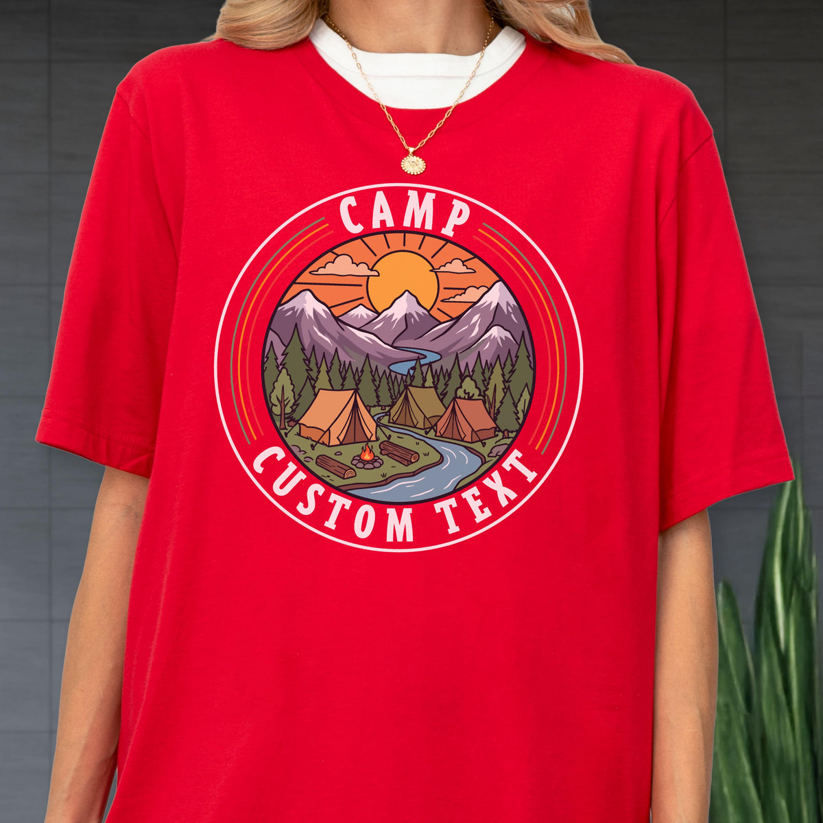 May include: A bright red t-shirt featuring a circular graphic with a mountain landscape, pine trees, a river, and tents. The text "CAMP" is arched at the top and "CUSTOM TEXT" is arched at the bottom of the circle, surrounding the outdoor scene.