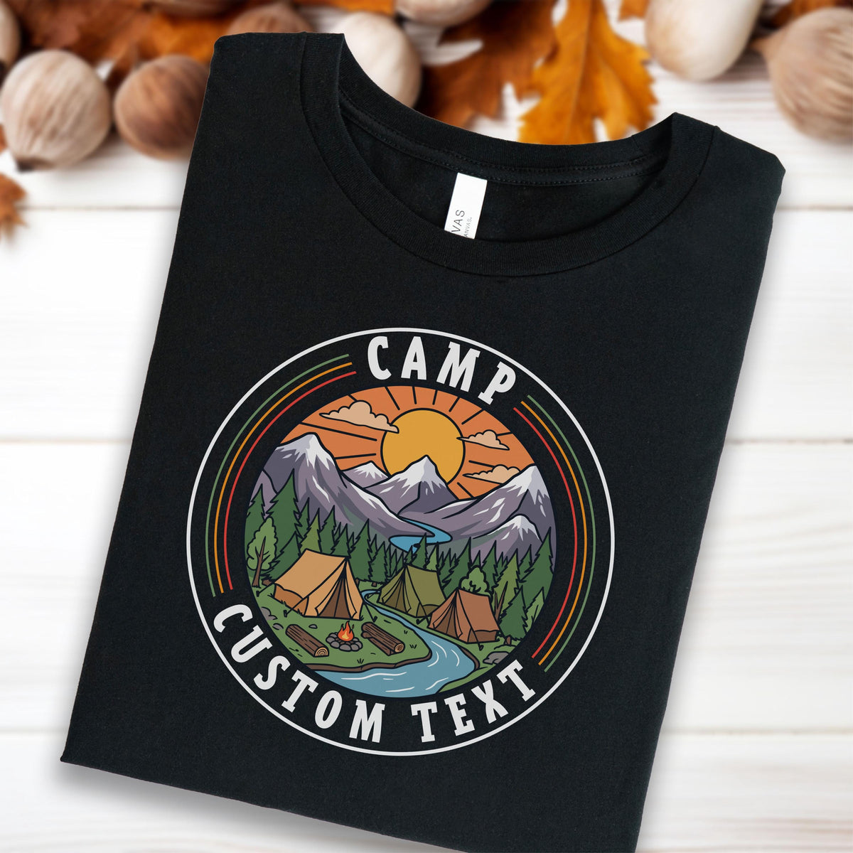 May include: A black t-shirt featuring a circular graphic with a mountain landscape, pine trees, a river, tents, and a campfire. The design includes a sun, clouds, and the text "CAMP" at the top and "CUSTOM TEXT" at the bottom in a white font.