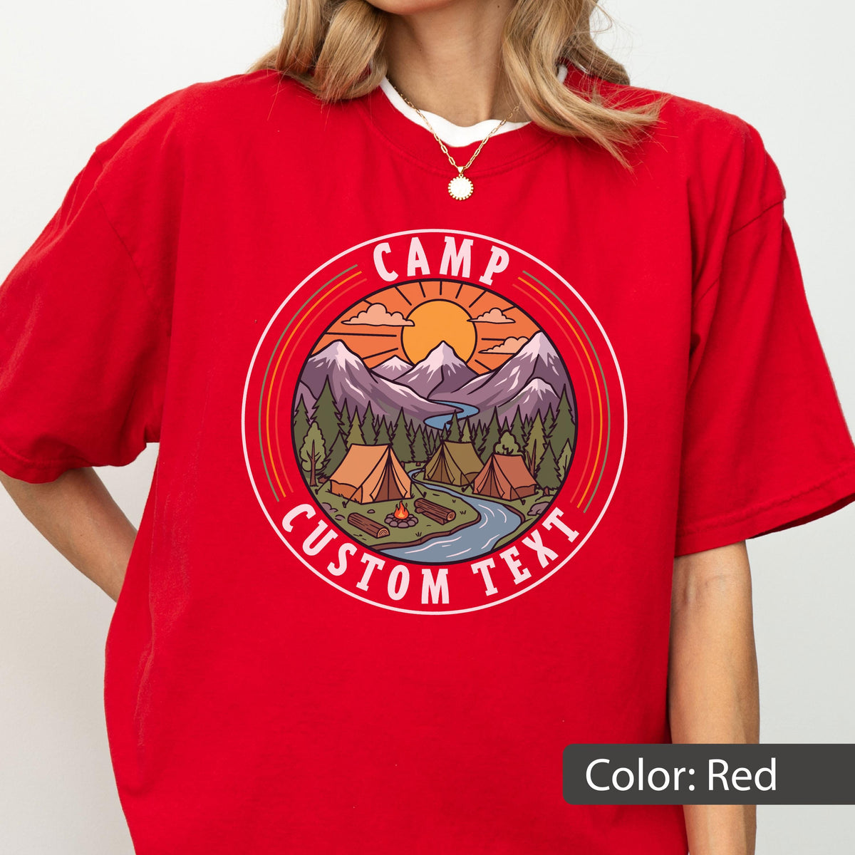 May include: A red t-shirt featuring a circular graphic of a mountain landscape with tents, a campfire, a river, and a sun. The text CAMP CUSTOM TEXT is printed in white within the circular border. The design uses orange, purple, and green tones.