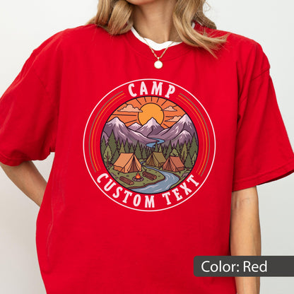 May include: A red t-shirt featuring a circular graphic of a mountain landscape with tents, a campfire, a river, and a sun. The text CAMP CUSTOM TEXT is printed in white within the circular border. The design uses orange, purple, and green tones.