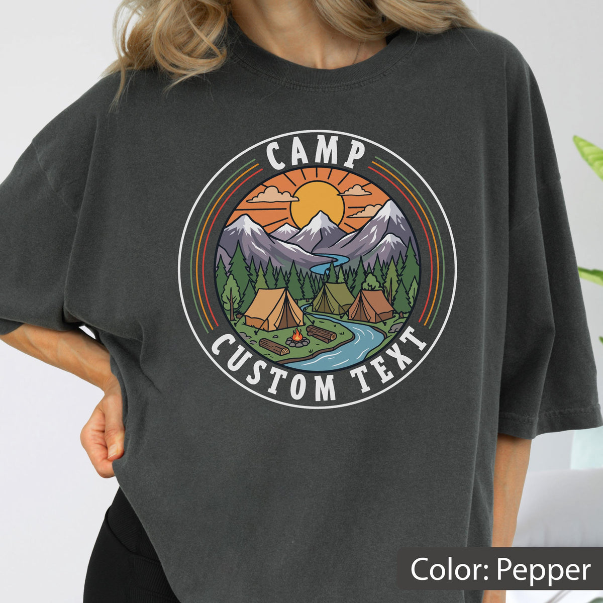 May include: A charcoal gray t-shirt featuring a circular graphic of a mountain landscape with a sun, pine trees, a river, and two tents. The text "CAMP" is arched at the top and "CUSTOM TEXT" is arched at the bottom in white capital letters.