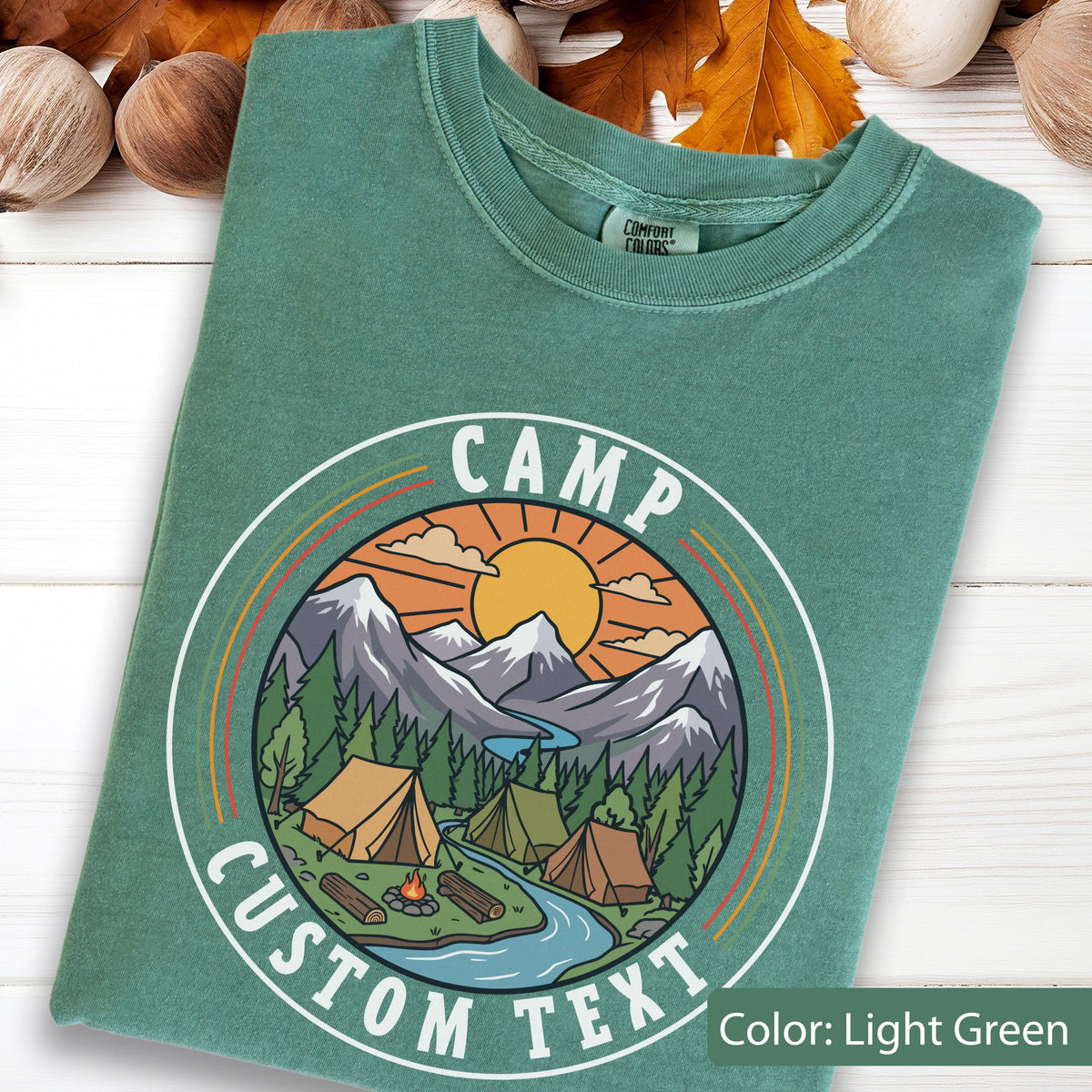 May include: A light green t-shirt featuring a circular graphic with the text "CAMP CUSTOM TEXT". The design depicts a sun, mountains, a river, pine trees, and three tents around a campfire. The shirt has a Comfort Colors brand label at the neck.