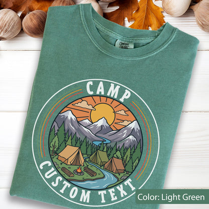 May include: A light green t-shirt featuring a circular graphic with the text "CAMP CUSTOM TEXT". The design depicts a sun, mountains, a river, pine trees, and three tents around a campfire. The shirt has a Comfort Colors brand label at the neck.
