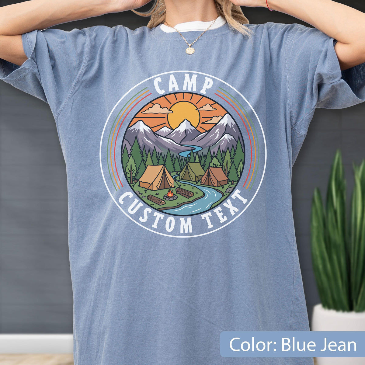 May include: A blue jean colored t-shirt featuring a circular graphic print of a mountain landscape with tents, a campfire, a river, and a sun. The text CAMP CUSTOM TEXT is printed in white within the circular border surrounding the illustration.