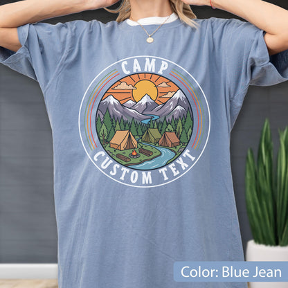 May include: A blue jean colored t-shirt featuring a circular graphic print of a mountain landscape with tents, a campfire, a river, and a sun. The text CAMP CUSTOM TEXT is printed in white within the circular border surrounding the illustration.