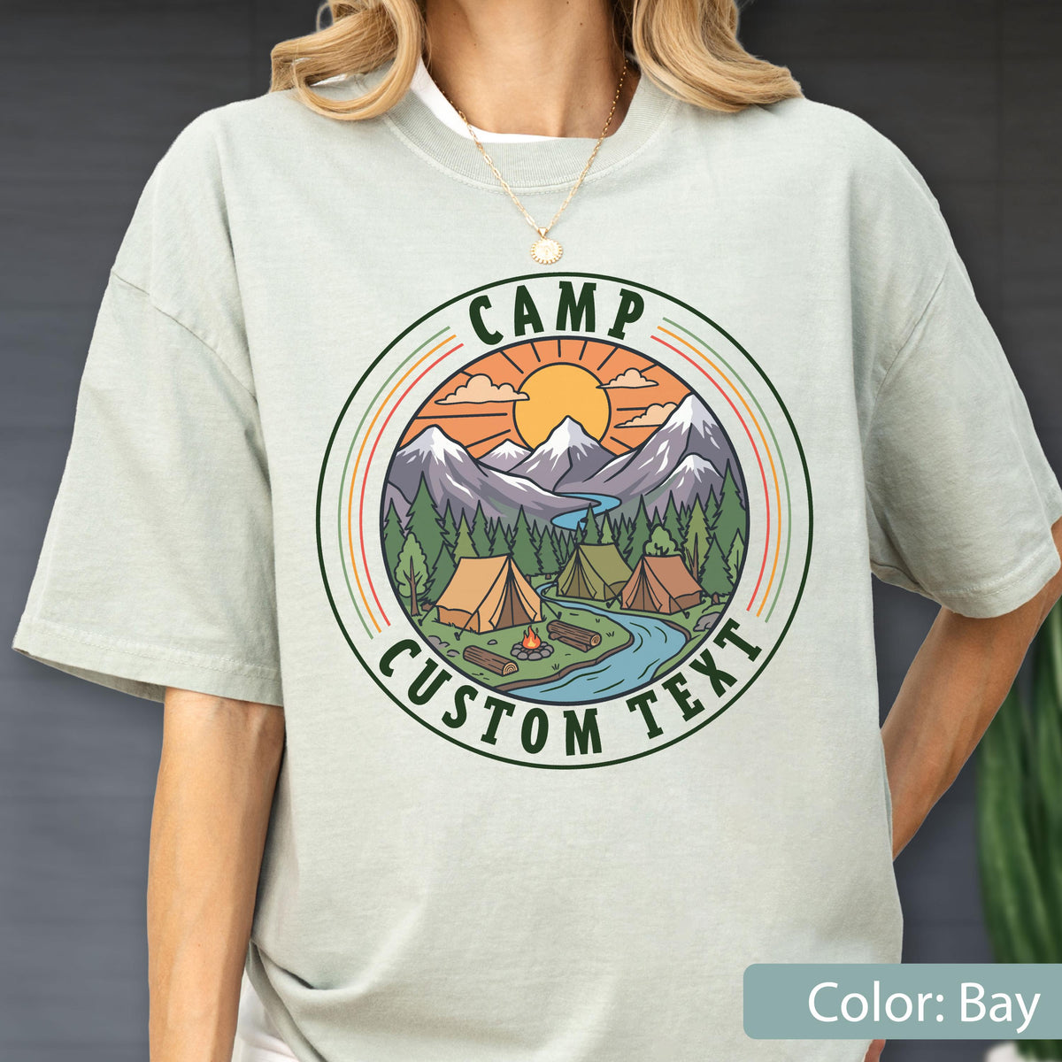 May include: A light sage green short-sleeve t-shirt featuring a circular graphic of a mountain landscape with tents, a river, and a campfire. The text CAMP CUSTOM TEXT is printed in dark green within the circular border around the illustration.
