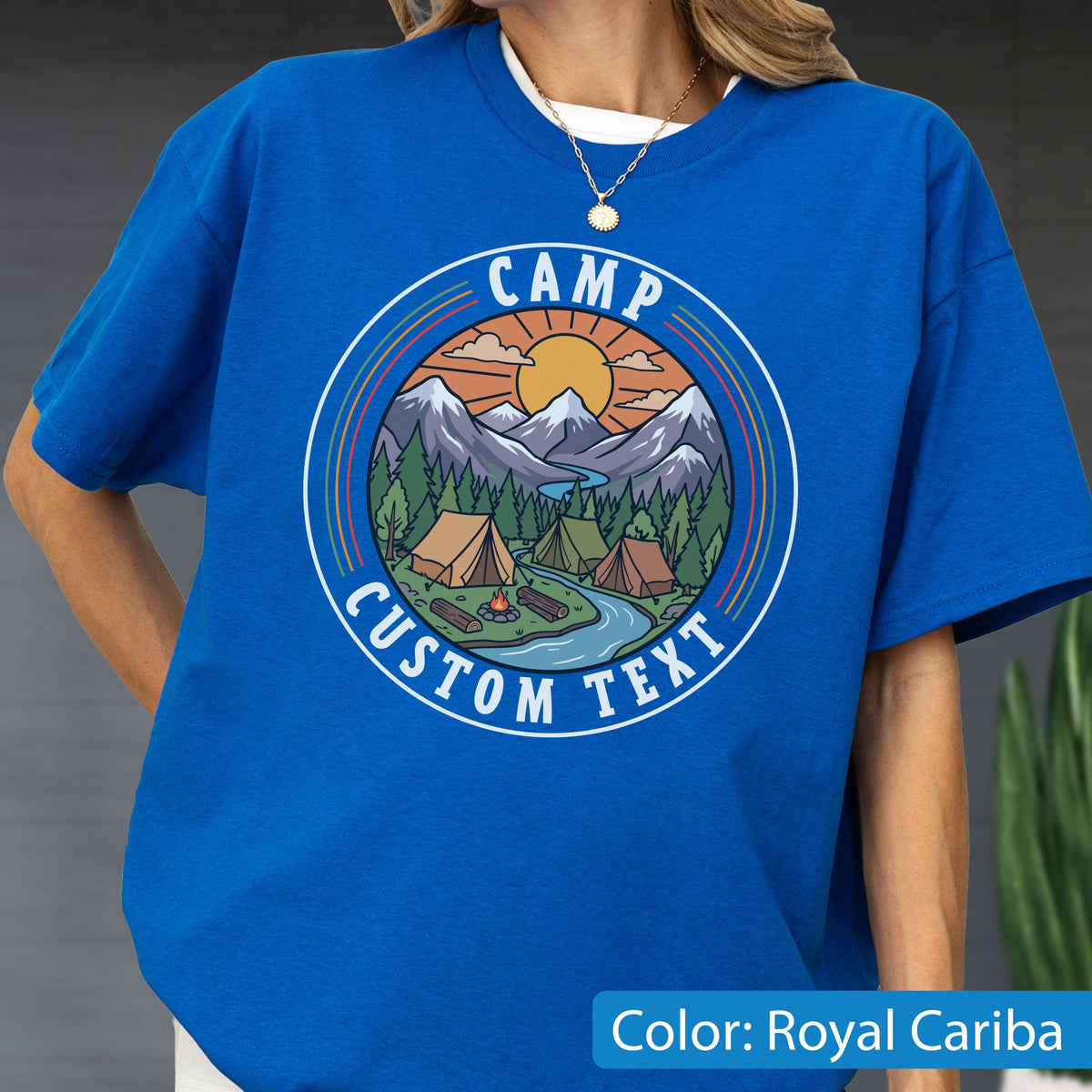 May include: A royal blue t-shirt featuring a circular graphic with a mountain landscape, pine trees, a river, and tents. The text CAMP is arched at the top and CUSTOM TEXT is arched at the bottom in white, sans-serif capital letters.