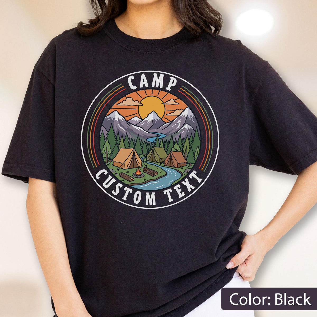 May include: A black t-shirt featuring a circular graphic with a mountain landscape, a river, pine trees, tents, and a campfire under a sun. The text CAMP and CUSTOM TEXT are printed in white within the circular border, which has orange and green rings.