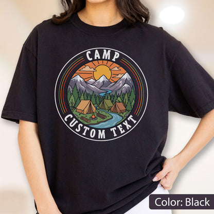 May include: A black t-shirt featuring a circular graphic with a mountain landscape, a river, pine trees, tents, and a campfire under a sun. The text CAMP and CUSTOM TEXT are printed in white within the circular border, which has orange and green rings.