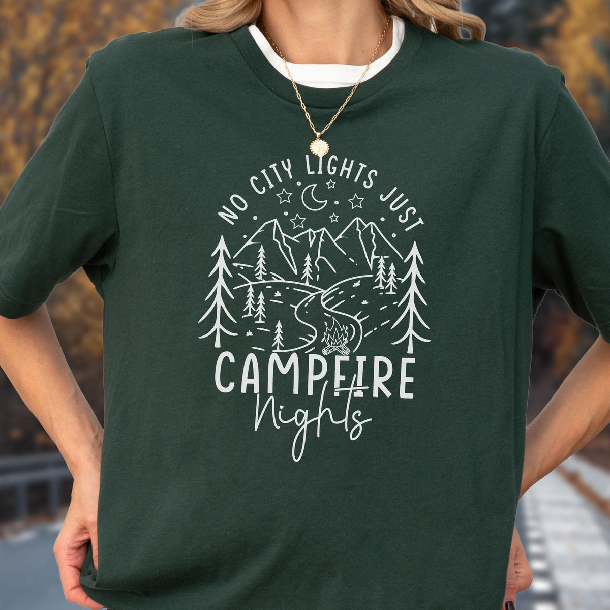 May include: A dark green short-sleeve t-shirt featuring a white graphic print of mountains, pine trees, a campfire, and stars. The text reads "NO CITY LIGHTS JUST CAMPFIRE Nights" in a mix of block and script lettering centered on the chest.