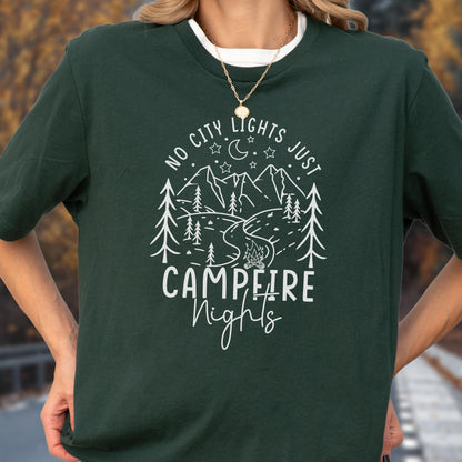 May include: A dark green short-sleeve t-shirt featuring a white graphic print of mountains, pine trees, a campfire, and stars. The text reads "NO CITY LIGHTS JUST CAMPFIRE Nights" in a mix of block and script lettering centered on the chest.