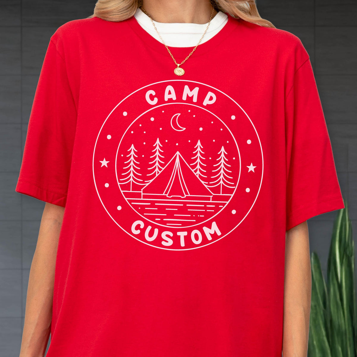 May include: A bright red short-sleeved t-shirt featuring a white circular graphic on the chest. The design includes the text CAMP CUSTOM, a central tent, pine trees, a crescent moon, and stars, all rendered in a clean, white line-art style.