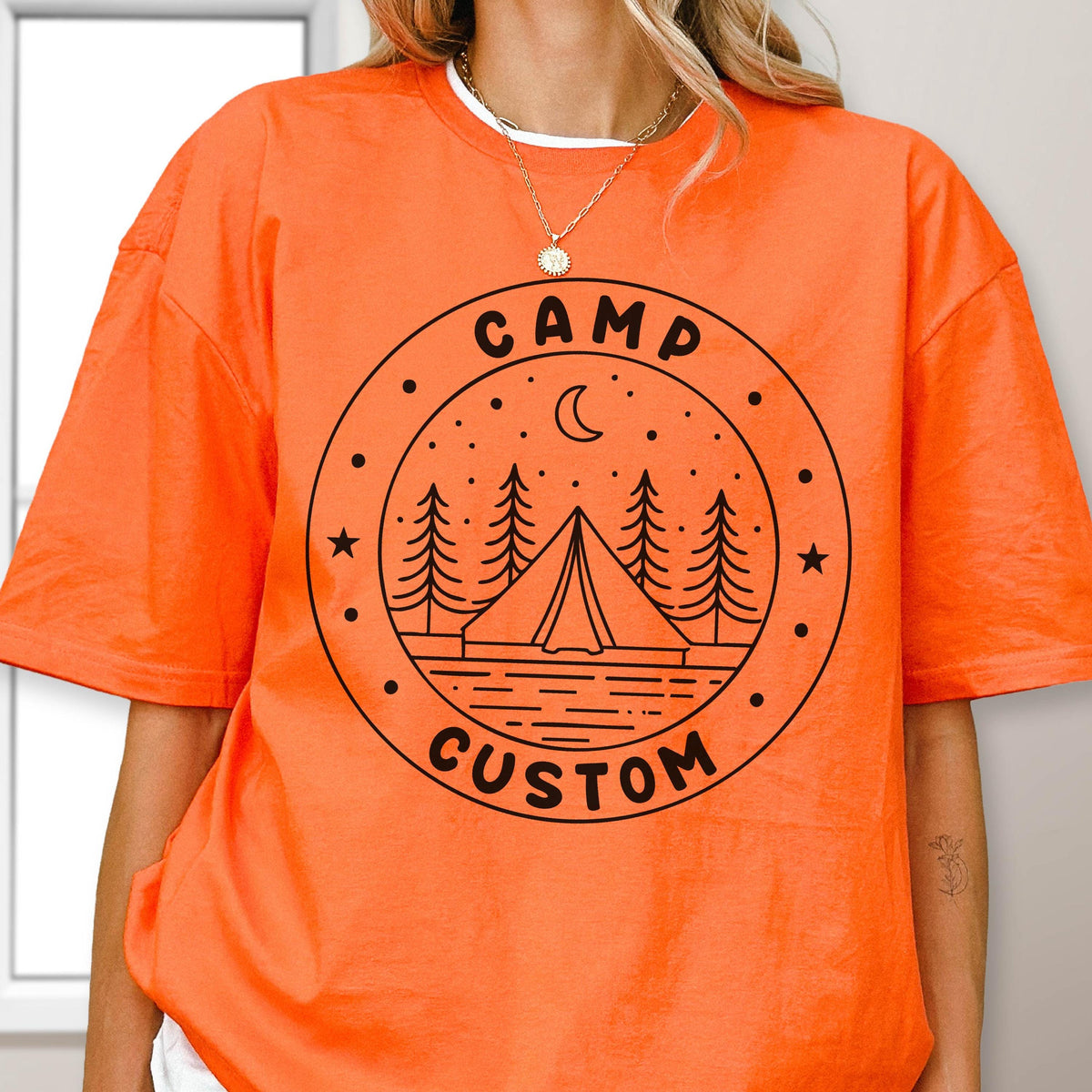 May include: A bright orange short-sleeved t-shirt featuring a black circular graphic on the chest. The design depicts a tent, pine trees, a crescent moon, and stars, with the text "CAMP CUSTOM" printed in a bold, sans-serif font along the border.