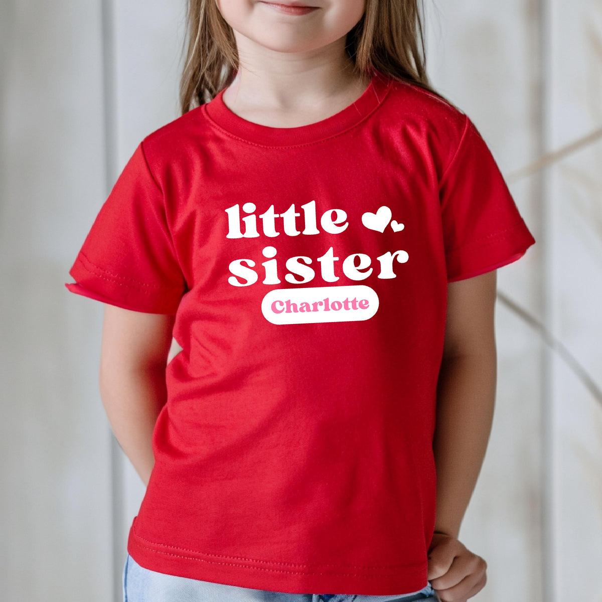 May include: A bright red short-sleeved t-shirt featuring the white text little sister in a bold, rounded font. Two small white hearts are positioned to the right of the text. Below, a white rectangular label displays the name Charlotte in pink.