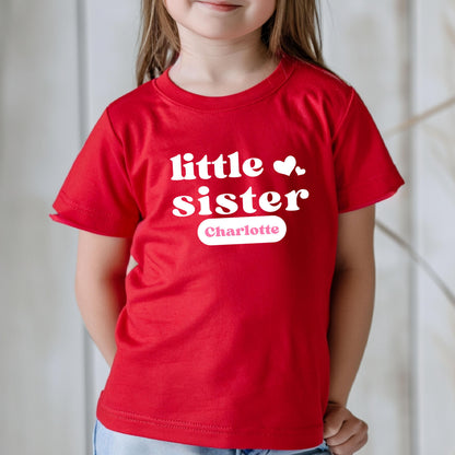 May include: A bright red short-sleeved t-shirt featuring the white text little sister in a bold, rounded font. Two small white hearts are positioned to the right of the text. Below, a white rectangular label displays the name Charlotte in pink.