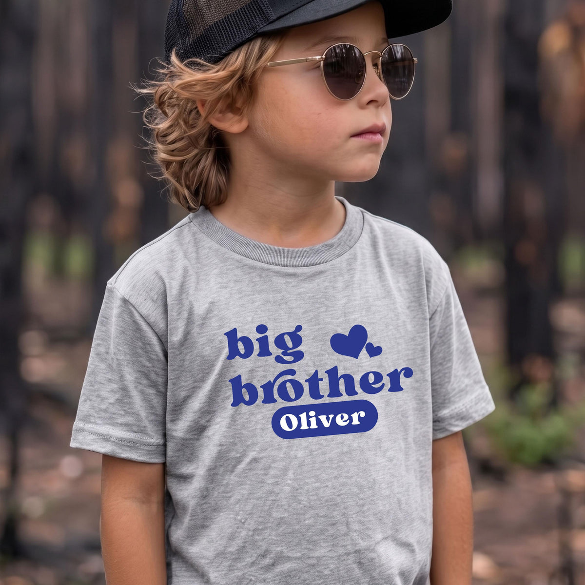 May include: A heather gray short-sleeved t-shirt featuring the blue text big brother and the name Oliver inside a blue rounded rectangle, accompanied by two blue heart icons. The shirt is worn with a black baseball cap and round sunglasses.
